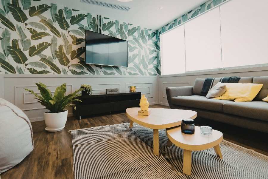 Un salon décoré avec un papier peint tropical à feuilles vertes, un canapé gris aux coussins jaunes et une table basse en bois pour donner du caractère à un salon.