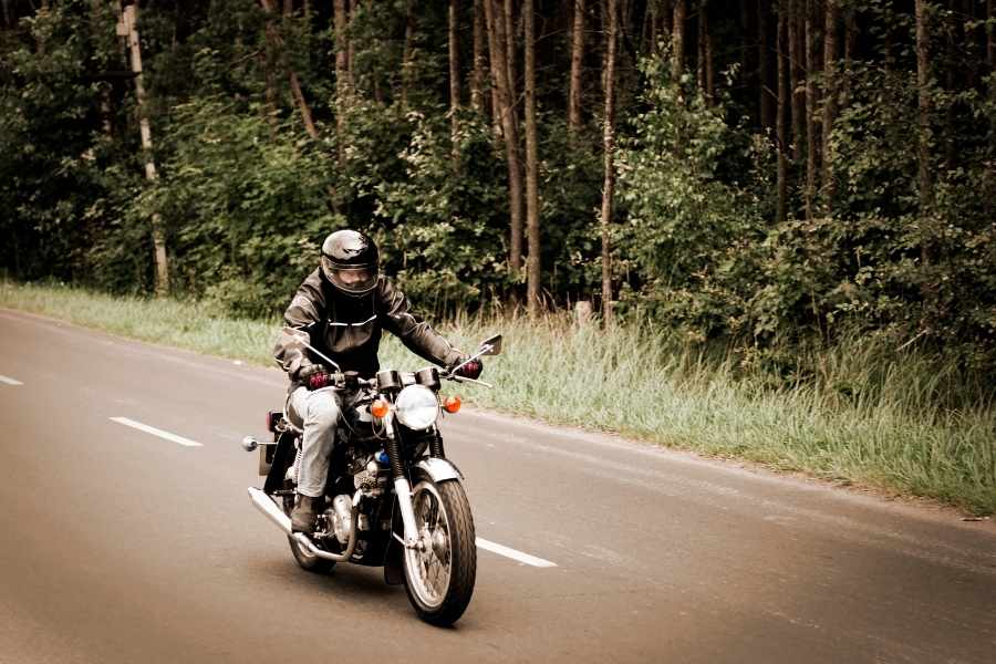 Un motard en veste noire et casque intégral roule sur une route forestière à bord d'une moto classique qui consomme moins grâce à une conduite souple et régulière.