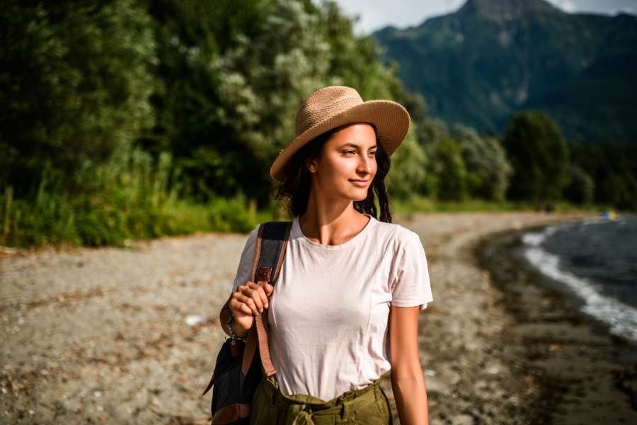 Une jeune femme souriante avec un sac à dos se promène au bord d'un lac naturel entouré de montagnes, incarnant les valeurs du tourisme durable.