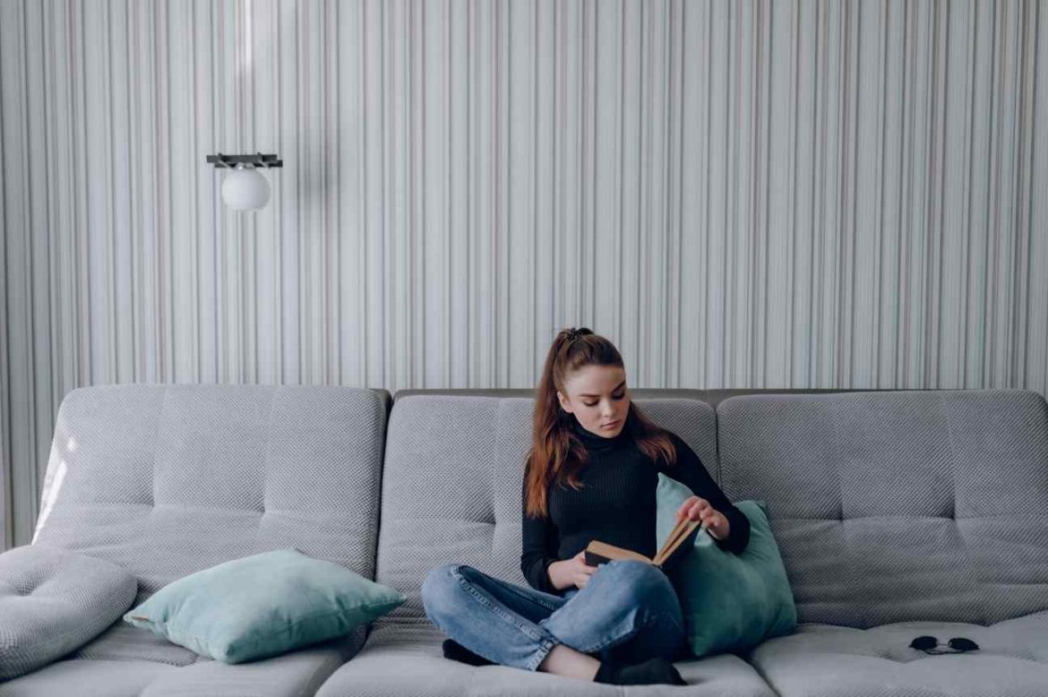 Une jeune femme lit un livre sur un canapé gris dans un salon rénové grâce à la pose chauffage mural hydraulique